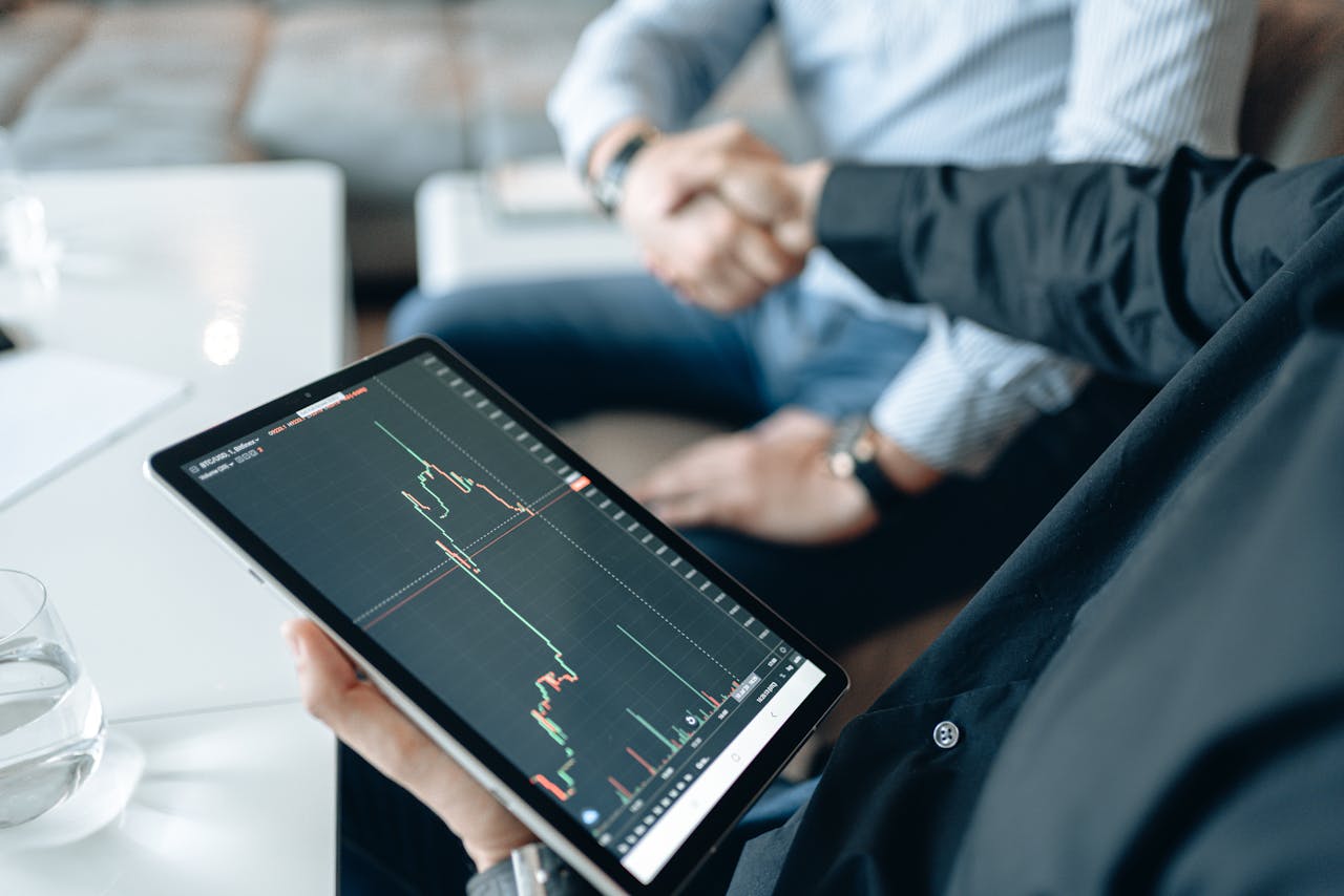 Two adults shaking hands with a stock market chart on a tablet, symbolizing a business deal.